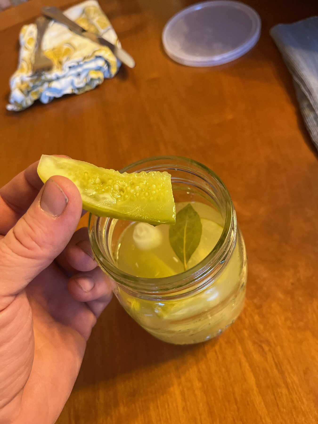 A homemade pickle held above an open mason jar with a bay leaf